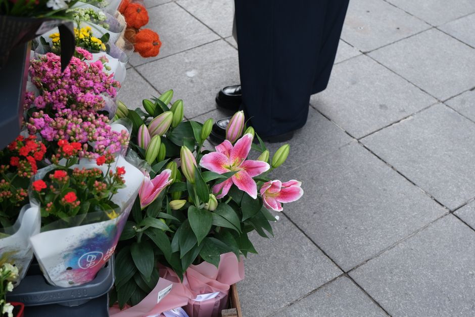 A floral display featuring a bouquet of pink lilies with large, open petals and prominent stamens, surrounded by closed lily buds and lush green leaves. The arrangement is placed on the pavement near