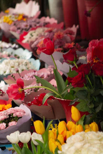 A vibrant display of fresh flowers arranged in various floral bouquets, including pink tulips, red and pink roses, yellow tulips, and white blossoms, all wrapped in decorative paper and placed on a ta