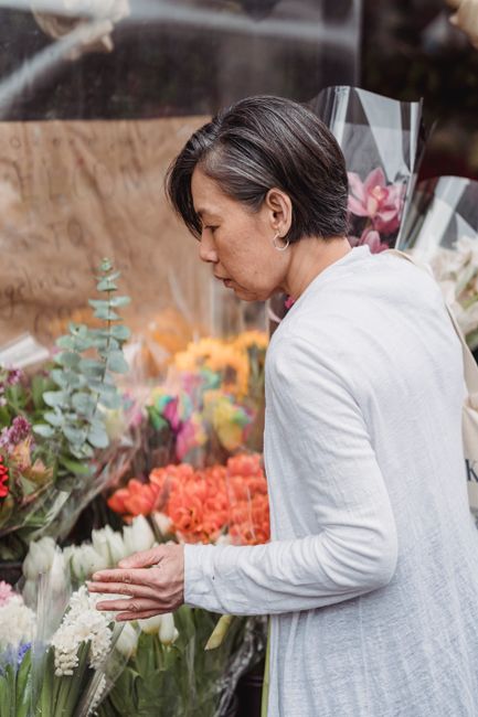 A woman with short dark hair dressed in a light grey blazer examining a colorful display of fresh flowers at a flower shop. The arrangement features a variety of blooms including orange roses, pink ca