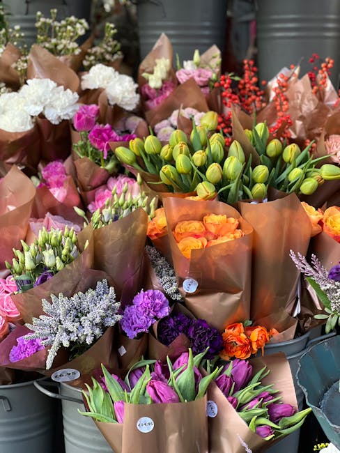 A vibrant display of freshly arranged flower bouquets featuring a variety of blooms such as pink and white carnations, orange roses, purple statice, lavender sprigs, and closed yellow tulips, all wrap