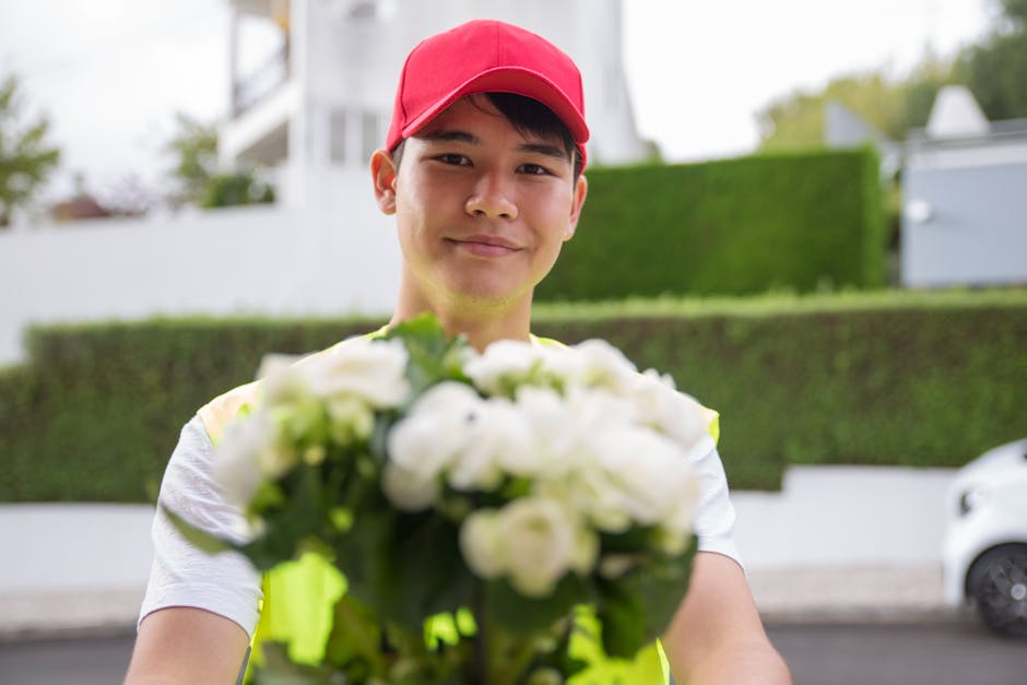 A delivery person wearing a red cap and a high-visibility vest is standing outdoors in a residential area, holding a bouquet of freshly cut white flowers with green leaves. The bouquet features variou